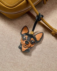Handmade Chihuahua leather bag charm clipped to a tan suede backpack — hand-cut French Alran leather and suede, embroidered eyes, contrast snout. STICKYZOO collectible.