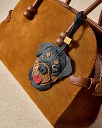 Handmade Rottweiler leather bag charm clipped to a tan suede handbag — full-grain French Alran goatskin, hand-stitched edges, 18K gold-plated hardware. STICKYZOO collectible.