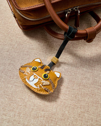 Ginger tabby hanging on a leather bag strap — three days of hand-work per piece. No machines. No shortcuts.