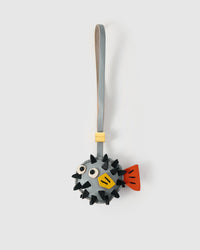 Pufferfish charm handmade from French Alran genuine goatskin — round gray body with real fiber spikes stitched in, stitched eyes, yellow mouth patch, orange side fin, gray leather strap. Three days by hand. Arrives in luxury gift packaging. Designed in Amsterdam.