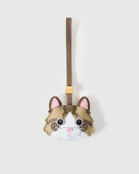 Pale tabby cat charm handmade from French Alran genuine goatskin — soft greige body, brown forehead stripes, pink inner ears, vivid blue eyes, white chin with pink nose, taupe leather strap. Three days by hand. Arrives in luxury gift packaging. Designed in Amsterdam.