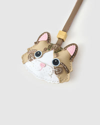 Both editions of the taupe-strap tabby cat charm side by side — leather strap loop for bags, or 18K gold-plated keychain clip with engraved STICKYZOO disc.