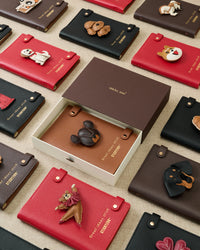 STICKYZOO notebook collection flat lay showing multiple leather charm designs in gift boxes, red, brown, and tan goatskin covers