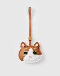 Patchwork cat charm handmade from French Alran genuine goatskin — caramel-tan body, white face mask and chin, pink inner ears, vivid green eyes, pink heart nose, tan leather strap. Three days by hand. Arrives in luxury gift packaging. Designed in Amsterdam.
