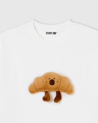 Close-up of plush croissant character with embroidered face and dangling legs on white cotton tee