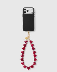 Black phone case with the ruby-red heart bead wrist strap looping below, on an 18K gold-plated hook and engraved disc.