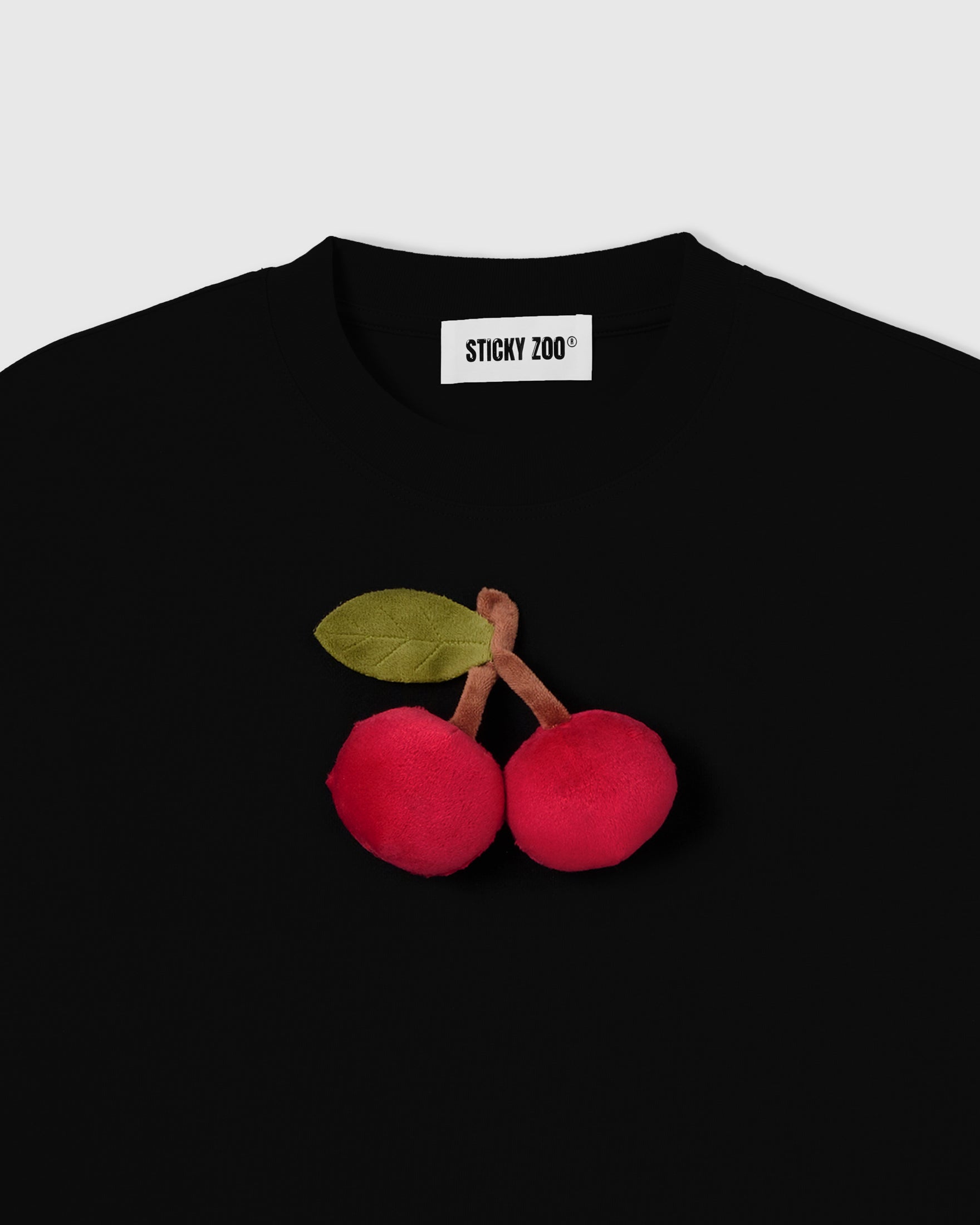 Close-up of plush cherry appliqué on black cotton showing velvety red bodies, brown stem, and green felt leaf with stitched veining