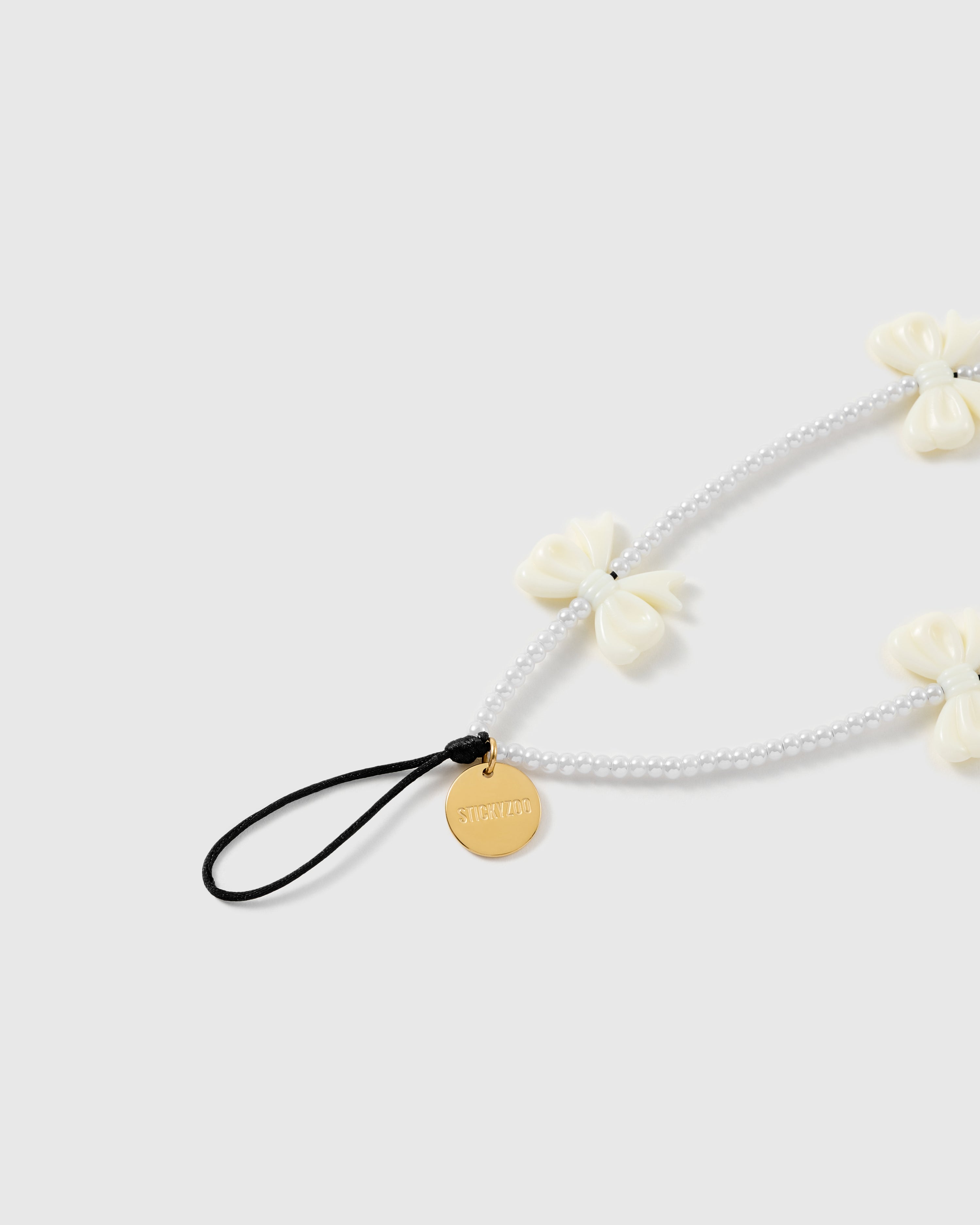 Close-up of the black stretch-cord attachment loop and gold-tone STICKYZOO disc, with white faux-pearl beads and a glossy ivory bow