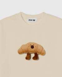 Close-up of plush croissant character with embroidered face and dangling legs on beige cotton tee