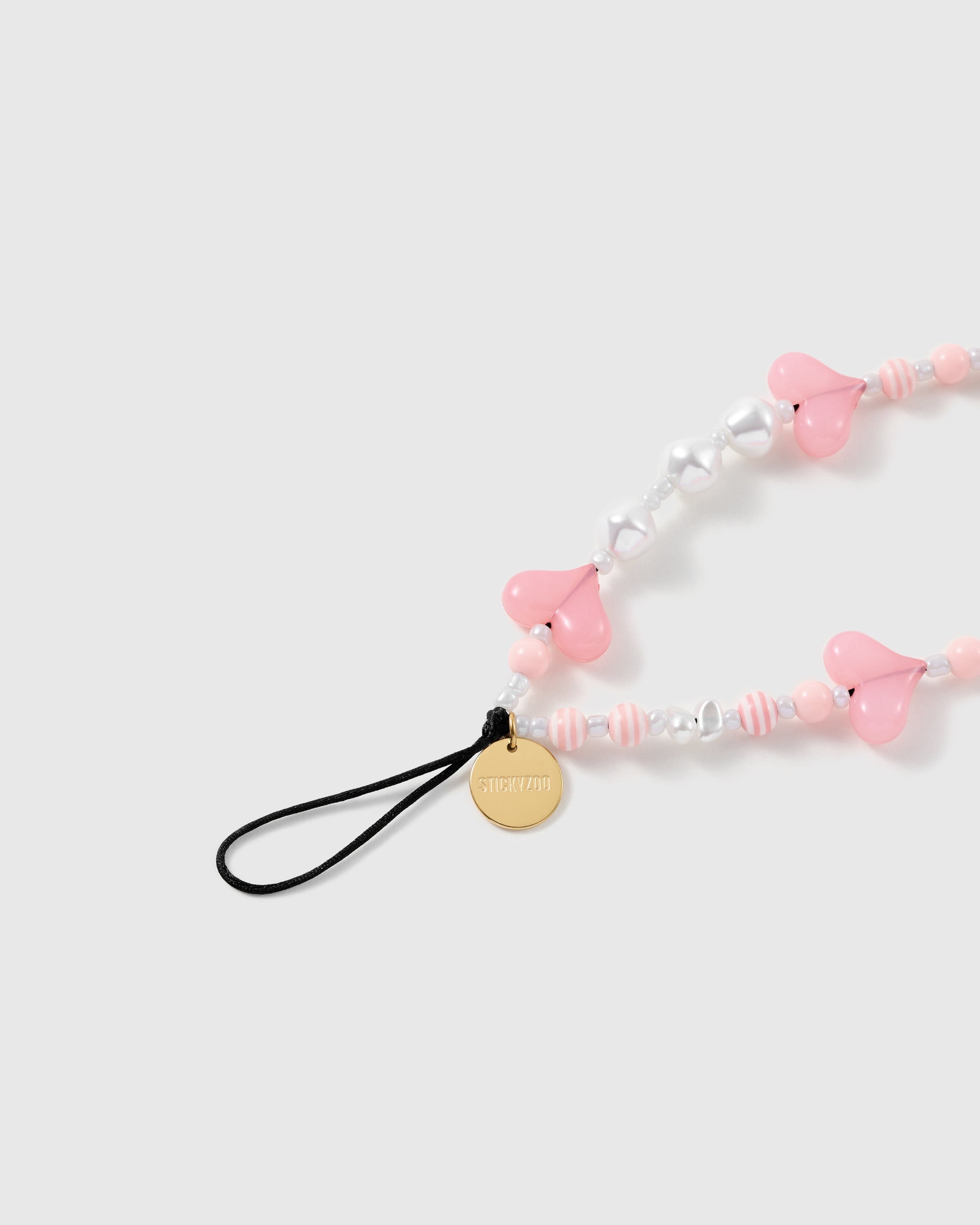 Close-up of the black stretch-cord loop and gold-tone engraved STICKYZOO disc beside pink striped, silver heart, and pearl-effect beads.
