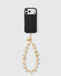 Black triple-camera phone case with the ivory heart bead wrist strap hanging in a loop from a gold hook and engraved gold disc.
