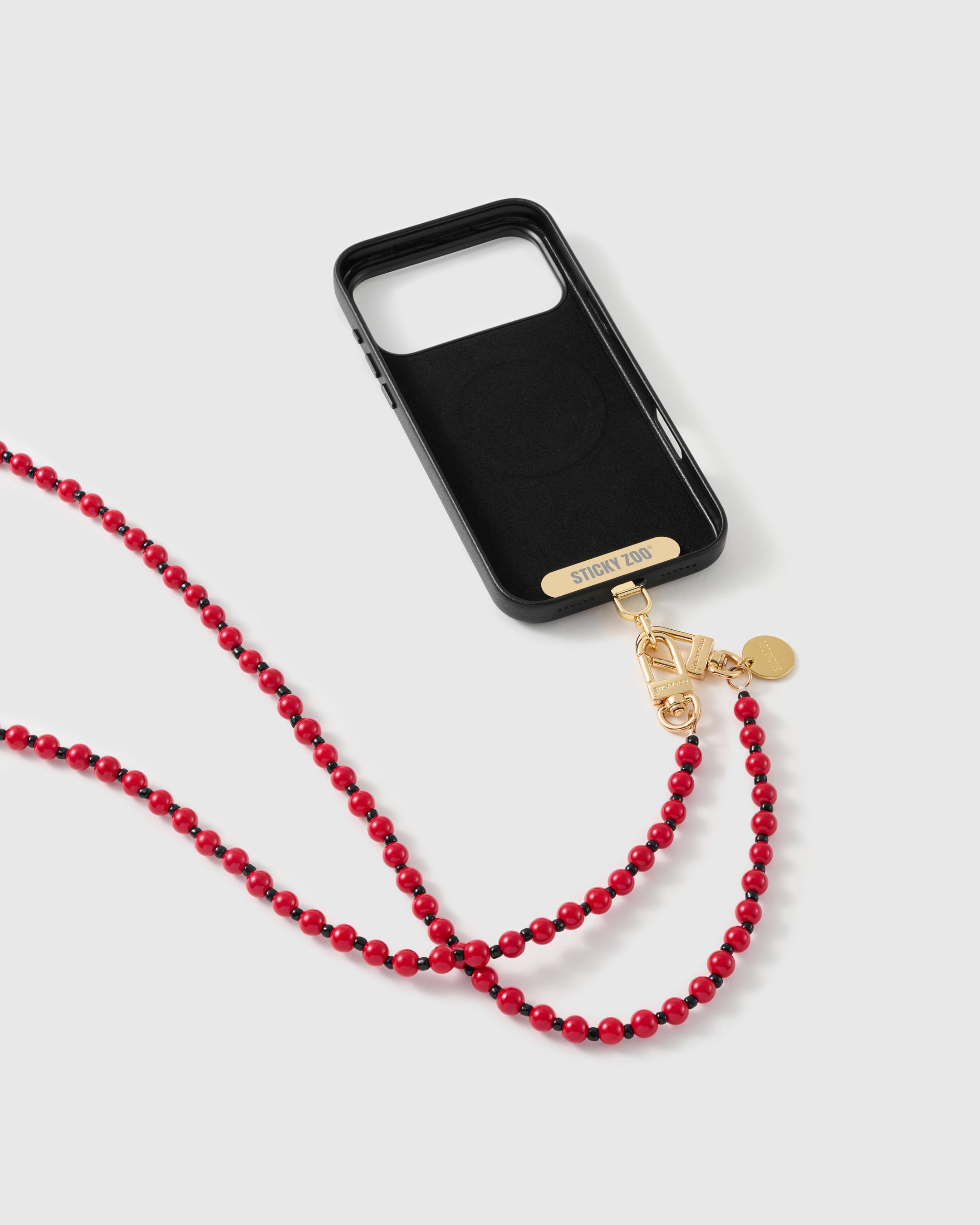Black phone case with a gold STICKYZOO bar and dual clasp holding the scarlet beaded strap, shown from the back on a white studio background.