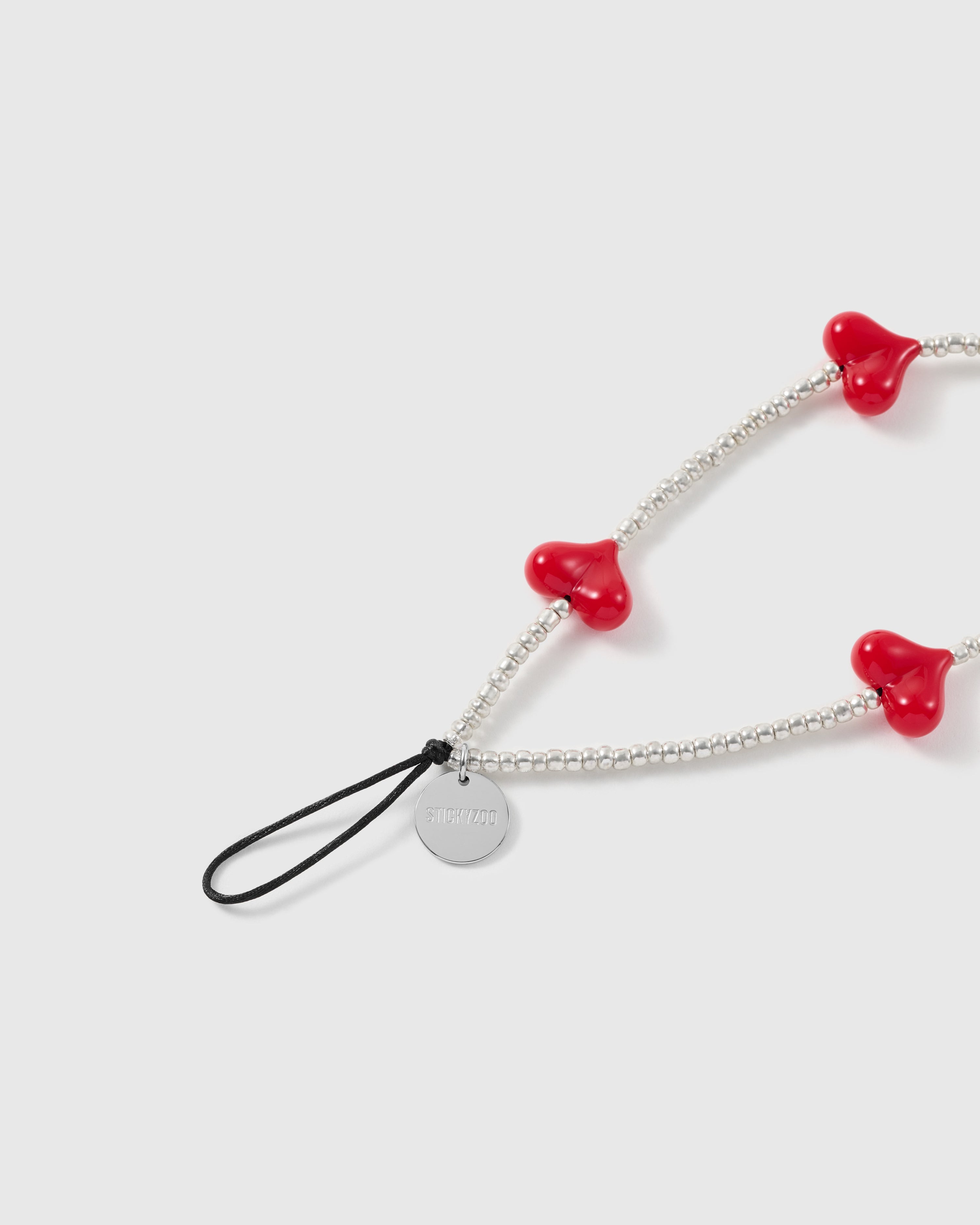 Close-up of the engraved silver-tone STICKYZOO disc and black stretch-cord loop, next to glossy red heart beads and silver seed beads.