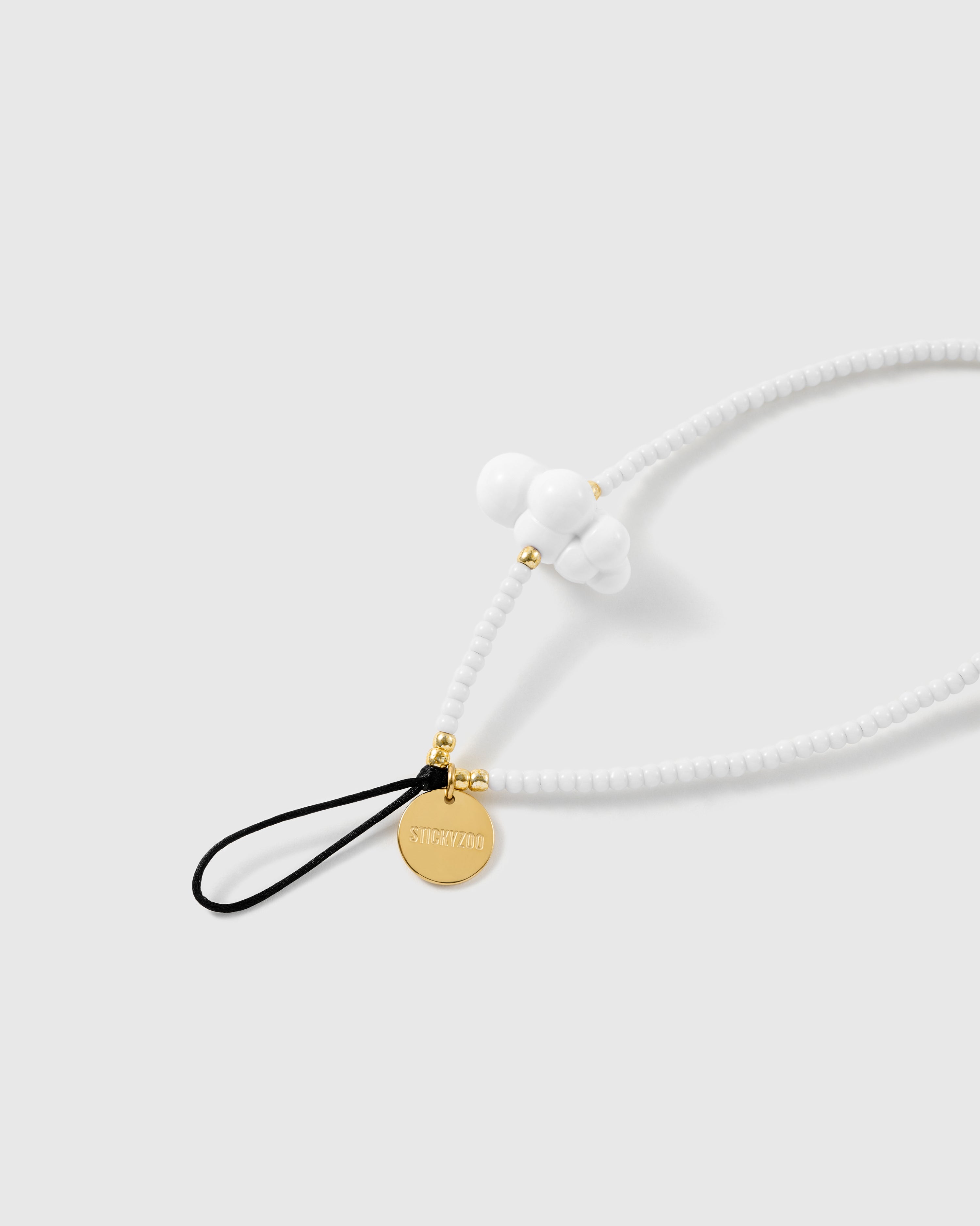 Close-up of the black stretch-cord loop and gold-tone STICKYZOO disc, with gold spacer beads and a white cloud bead on white seed beads.