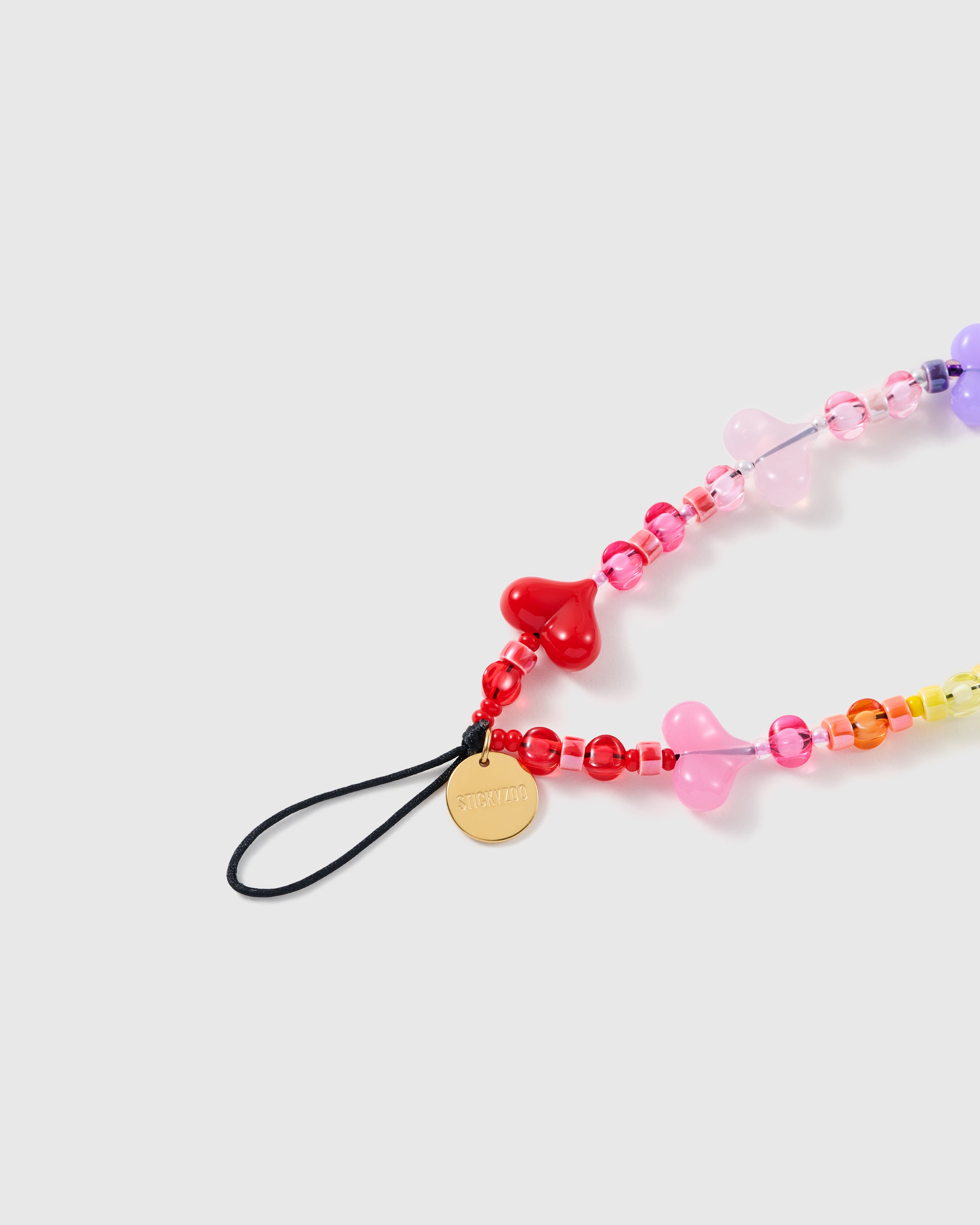 Close-up of the gold-tone STICKYZOO disc and black stretch-cord loop beside a glossy red jelly heart and clear rainbow beads.