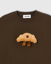 Close-up of plush croissant applique on brown cotton showing 3D stuffed texture with embroidered face and plush boots