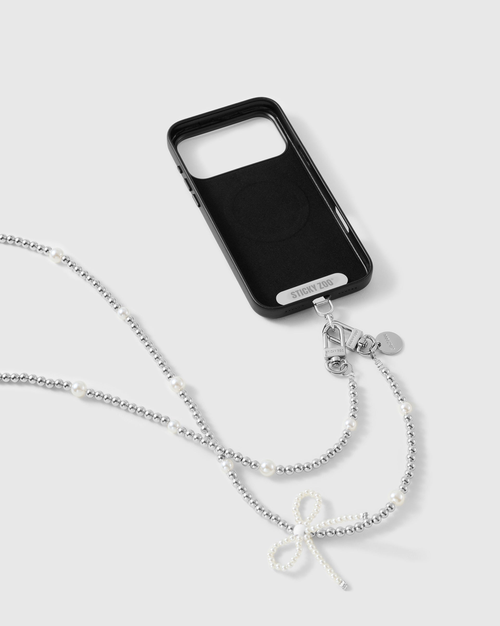 STICKYZOO strap clipped to a black phone case by its dual silver-tone clasp, beaded strand and faux-pearl bow trailing below.