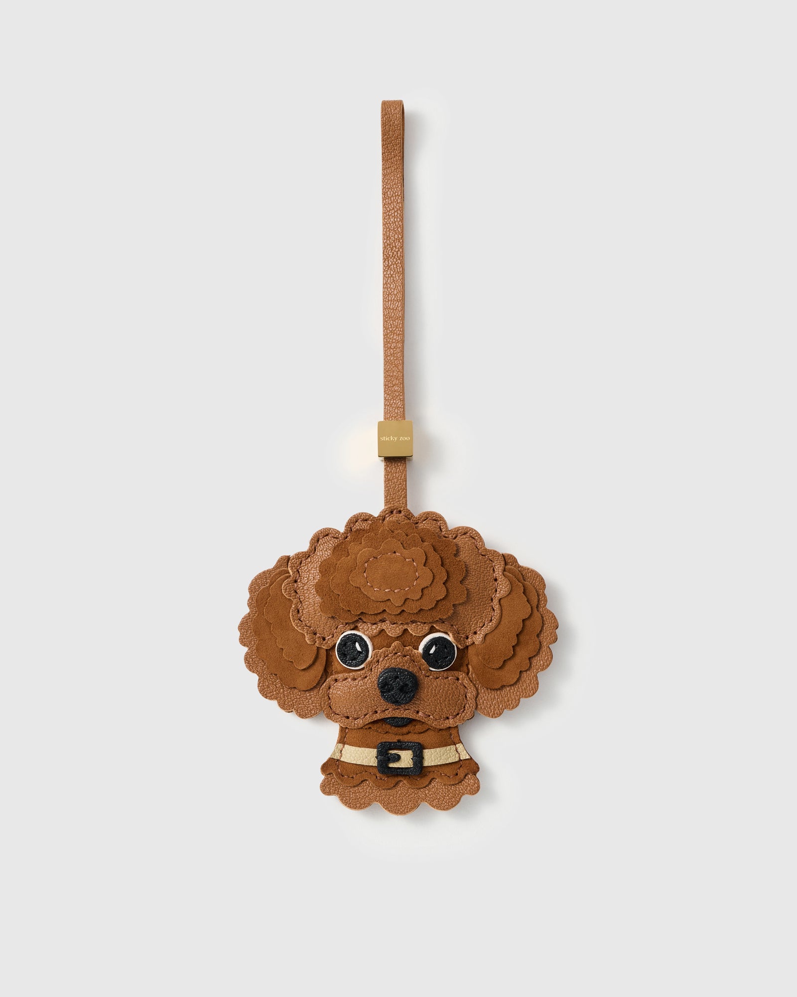 {{Alcantara × French leather.}} The Poodle Charm — exclusive drop from STICKYZOO. Hand-cut French Alran goatskin and Italian Alcantara, warm cognac tones, layered scalloped curls, gold collar with black buckle, cognac leather strap. Seven days per piece. Designed in Amsterdam.