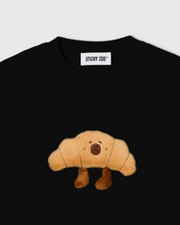 Close-up of plush croissant character with embroidered face and dangling legs on black cotton tee