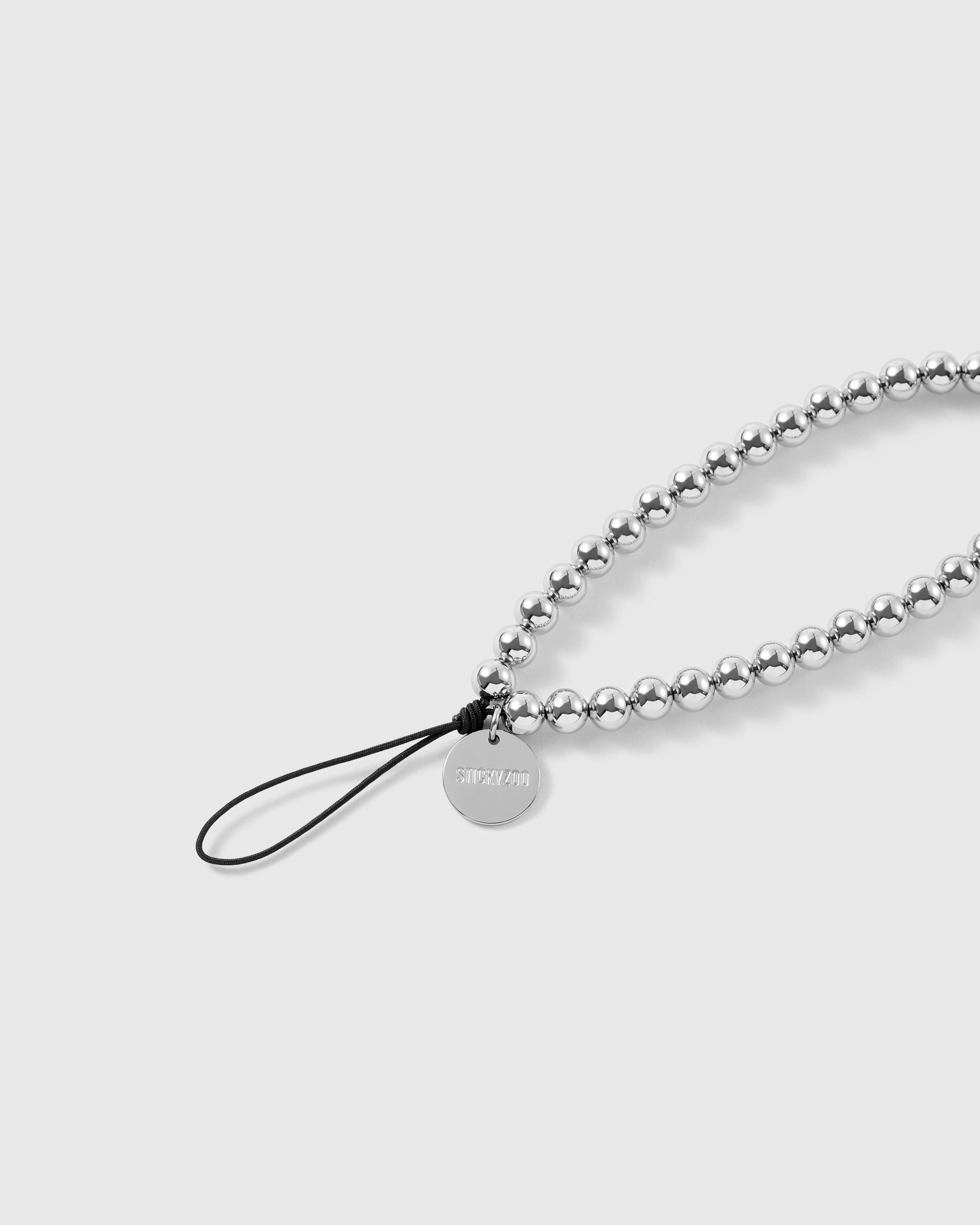 Close-up of the black stretch-cord loop knotted to an engraved silver-tone STICKYZOO disc beside smooth silver ball beads