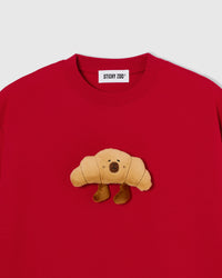 Close-up of hand-stitched plush croissant on red 230gsm cotton showing stuffed texture and embroidered face