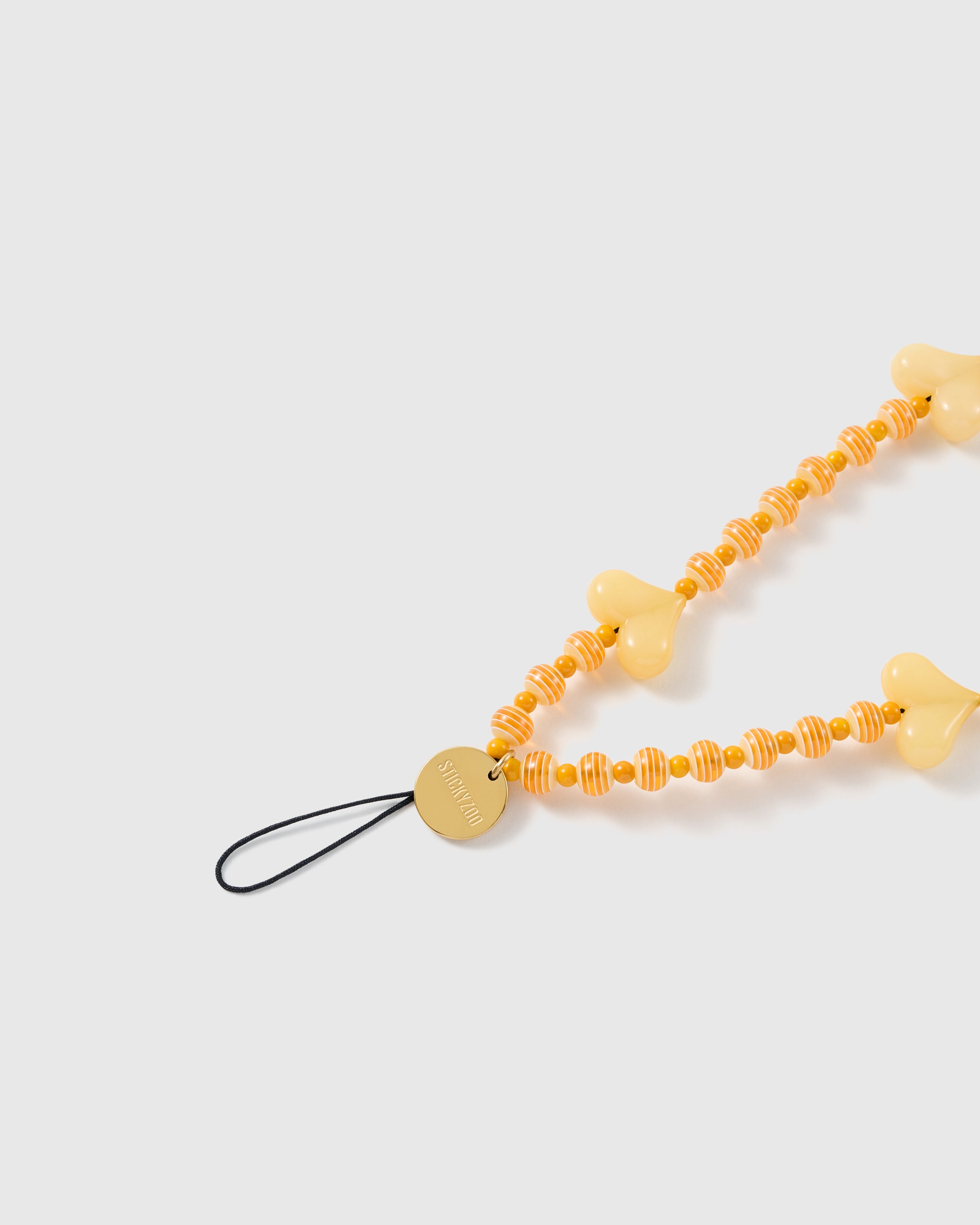 Close-up of the STICKYZOO gold-tone engraved disc and black stretch-cord loop beside orange-and-cream striped beads and a butter-yellow heart.