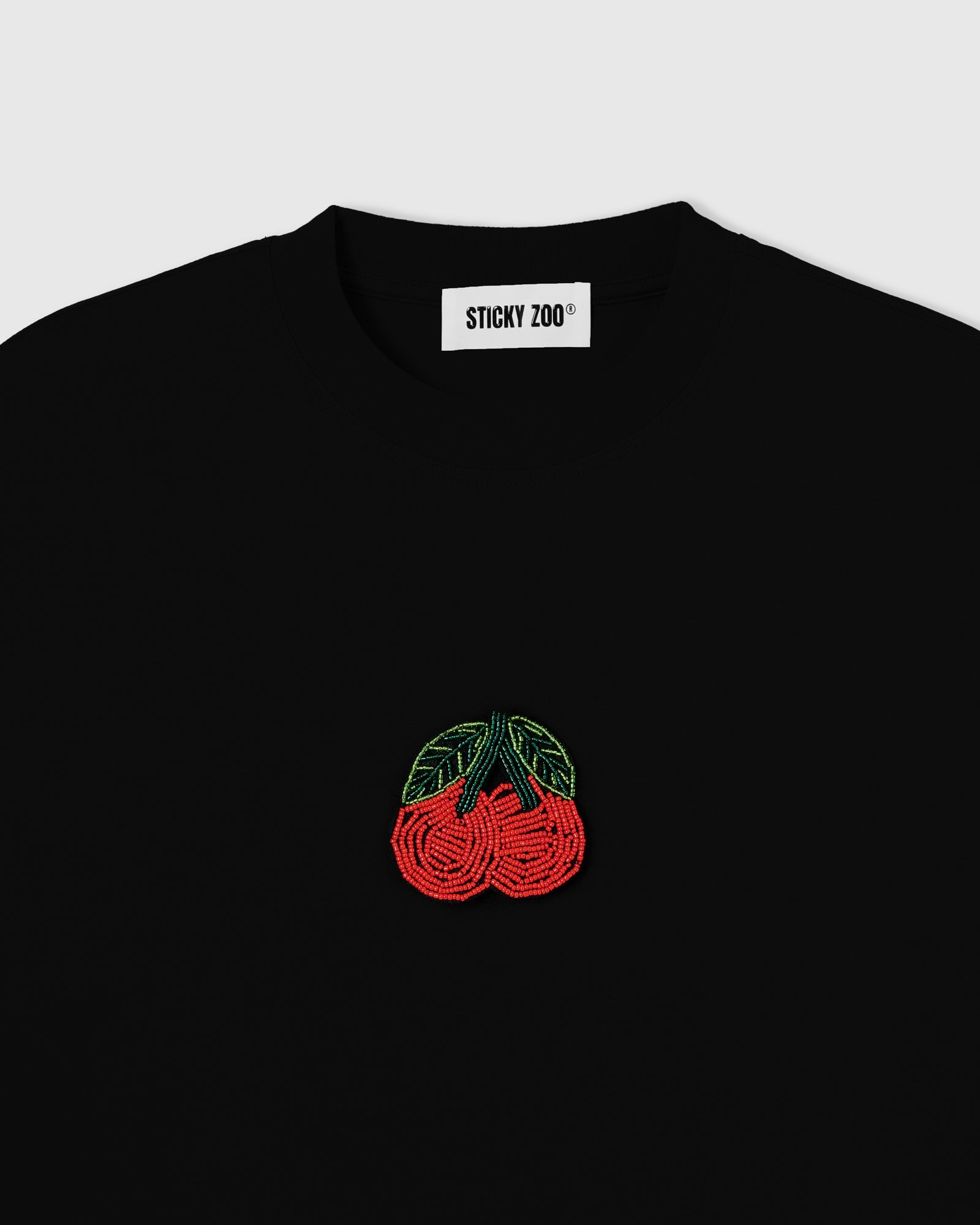 Close-up of the hand-beaded cherry appliqué and woven STICKYZOO neck label on the black 230gsm cotton tee, showing glass seed beads set by hand