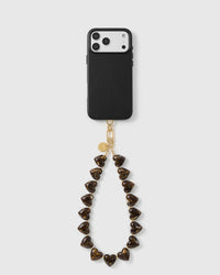 Tortoiseshell Heart wrist strap looped below a black phone case, with marbled amber heart beads, a gold-tone hook and engraved disc.