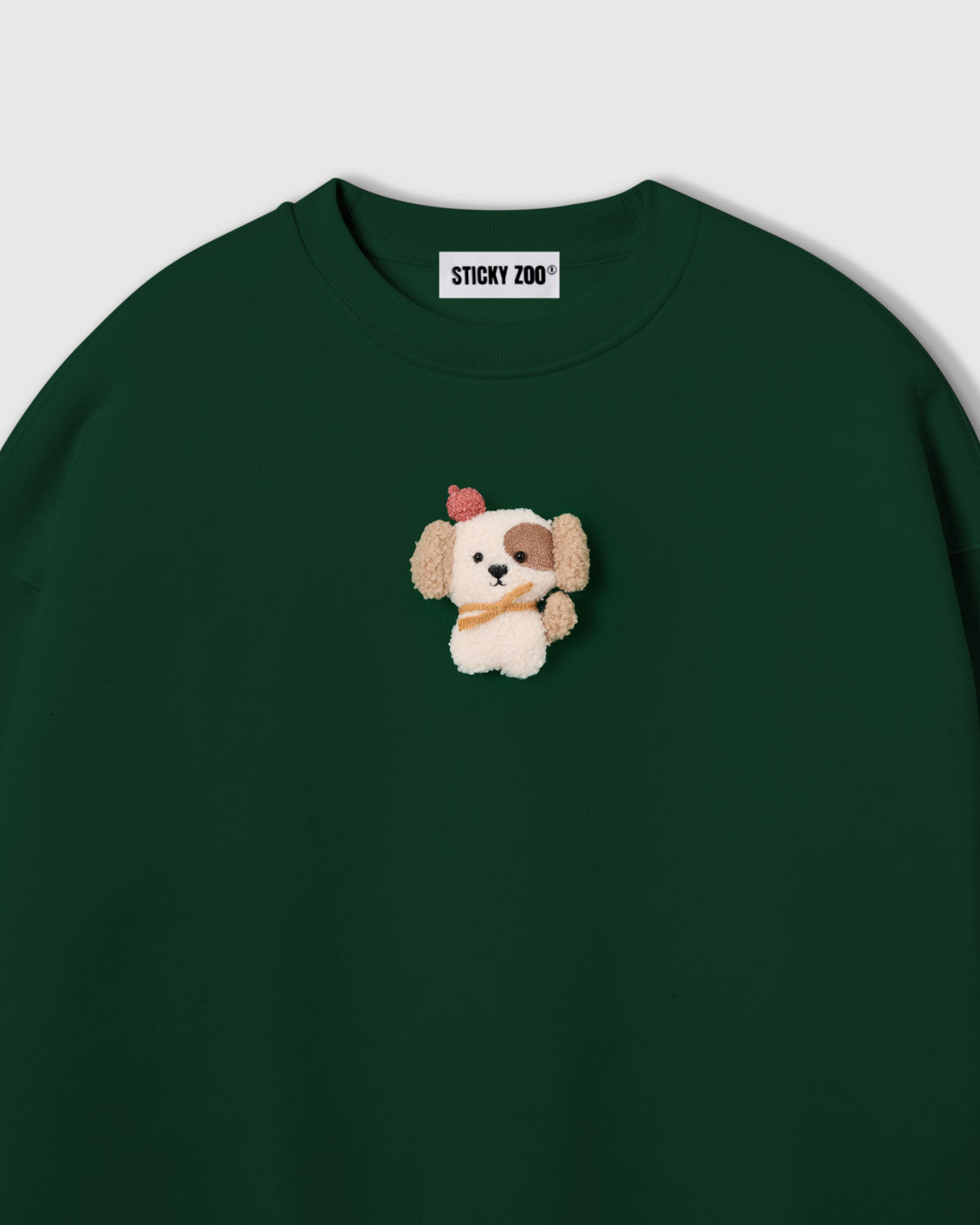 {{Not printed — stitched}} Close-up of hand-stitched plush puppy on green cotton showing cream sherpa body, floppy ears, and golden scarf detail
