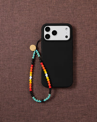 Multicolour beaded phone strap looped onto a black phone case on brown linen, gold disc and opaque pony beads in turquoise, red, orange and yellow.