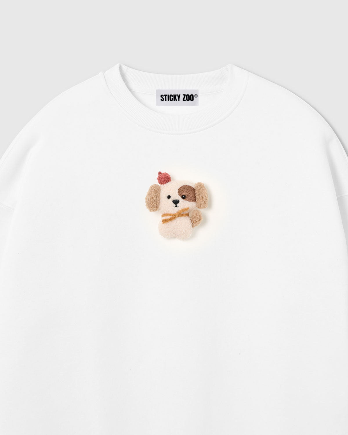 {{Not printed — stitched}} Close-up of hand-stitched plush puppy on white cotton showing cream sherpa body and tonal texture contrast