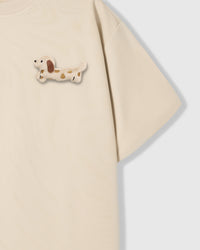 Close-up of spotted dachshund plush on shoulder area of beige tee showing plush detail