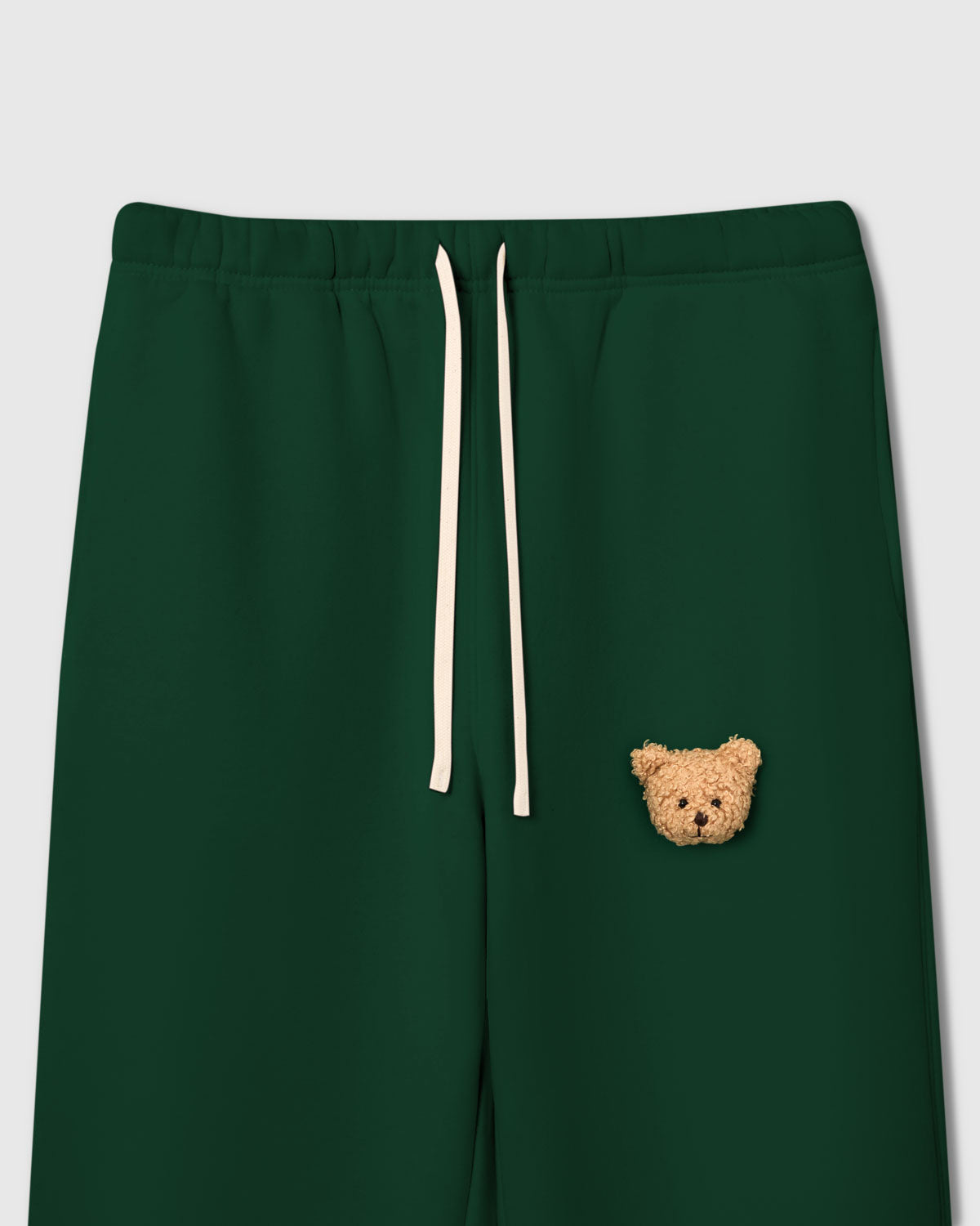 {{Not printed — stitched}} Close-up of elastic waistband and plush teddy bear face applique on green 350gsm cotton showing cream drawstring and sherpa texture
