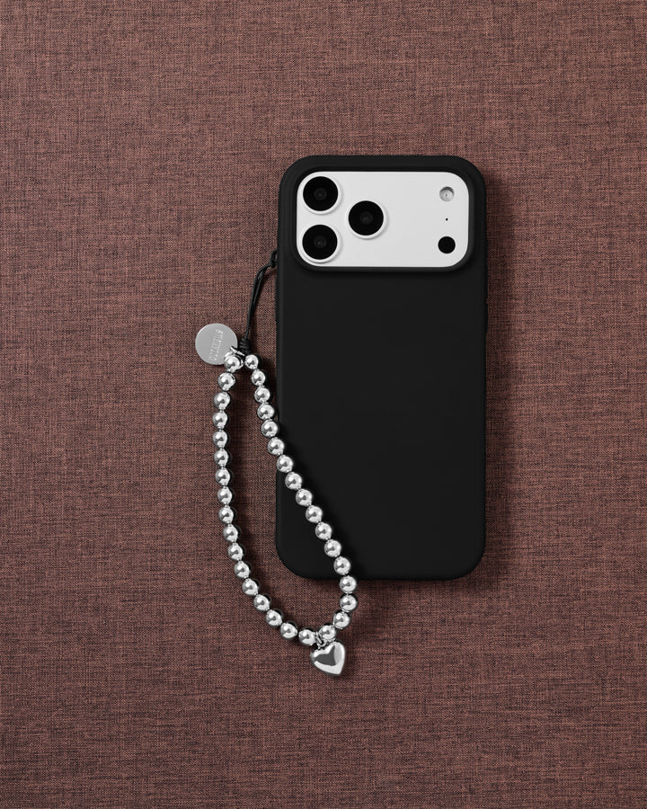 STICKYZOO beaded phone strap draped beside a black phone case on woven mauve fabric, silver ball beads ending in a puffy heart drop