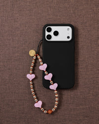 STICKYZOO Hazel Heart strap looped on a black phone case over brown linen, striped brown-and-cream beads and a rust-brown round at the base