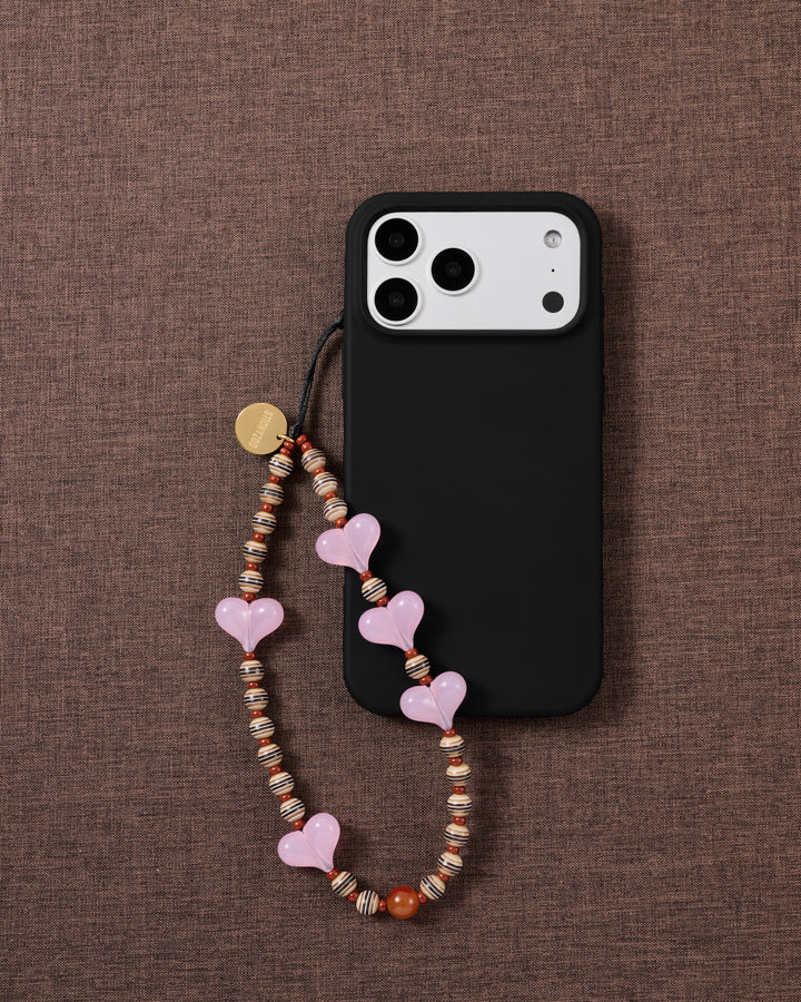 STICKYZOO Hazel Heart strap looped on a black phone case over brown linen, striped brown-and-cream beads and a rust-brown round at the base
