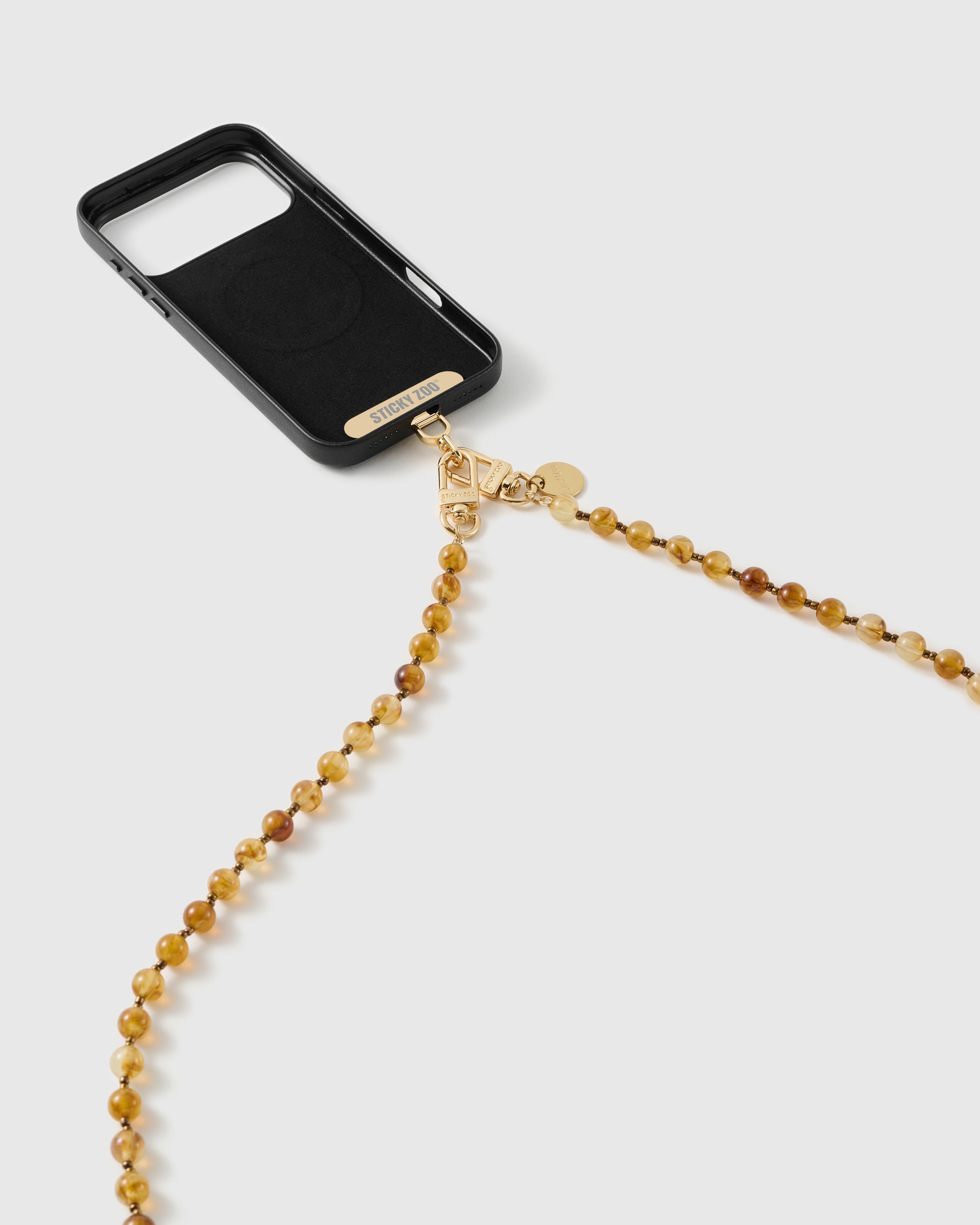 Amber resin bead strap clipped to a black phone case via gold-tone dual clasps and a round engraved disc, strap trailing below.