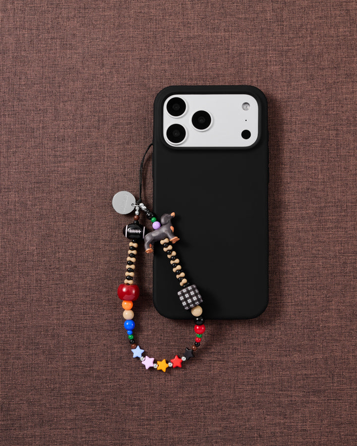 The beaded dachshund phone strap looped on a black phone case over brown linen, showing rainbow beads and multicolour stars.