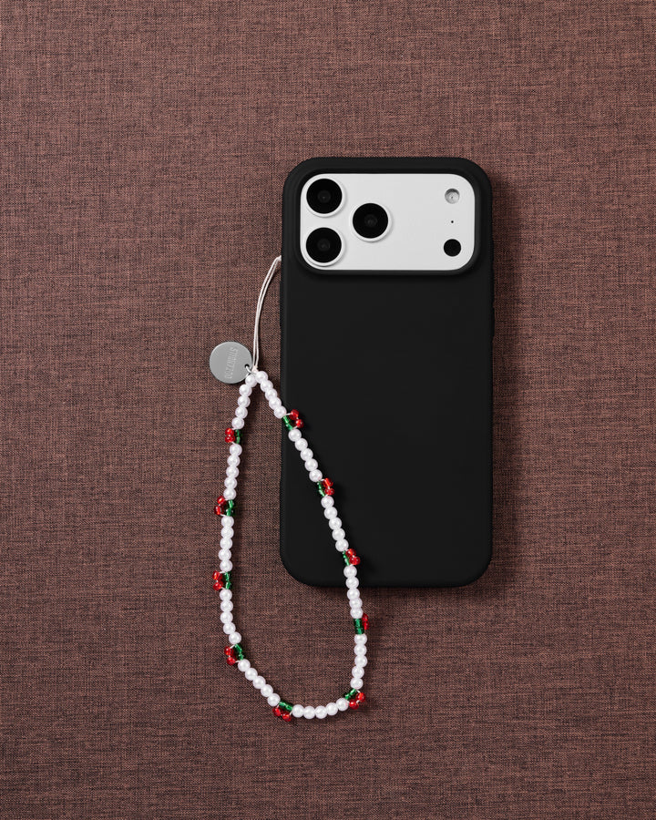 Holly-berry faux-pearl phone strap looped onto a black phone case on brown fabric, showing red-and-green berry beads and the silver disc.