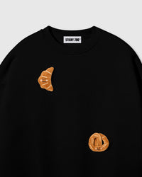 Close-up of crescent plush croissant with smiley face on black cotton showing golden-brown layered texture