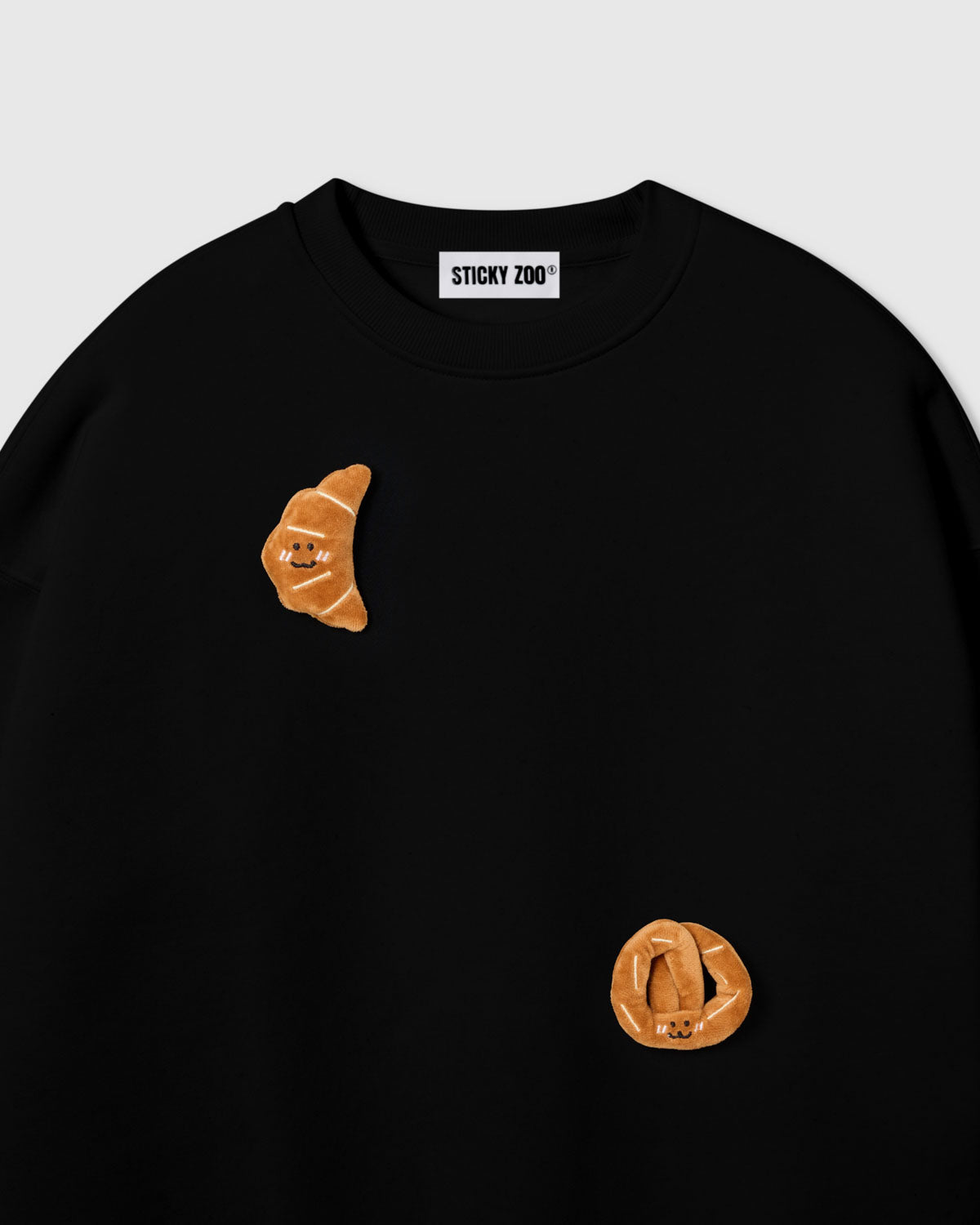 {{Not printed — stitched}} Close-up of crescent plush croissant with smiley face on black cotton showing golden-brown layered texture