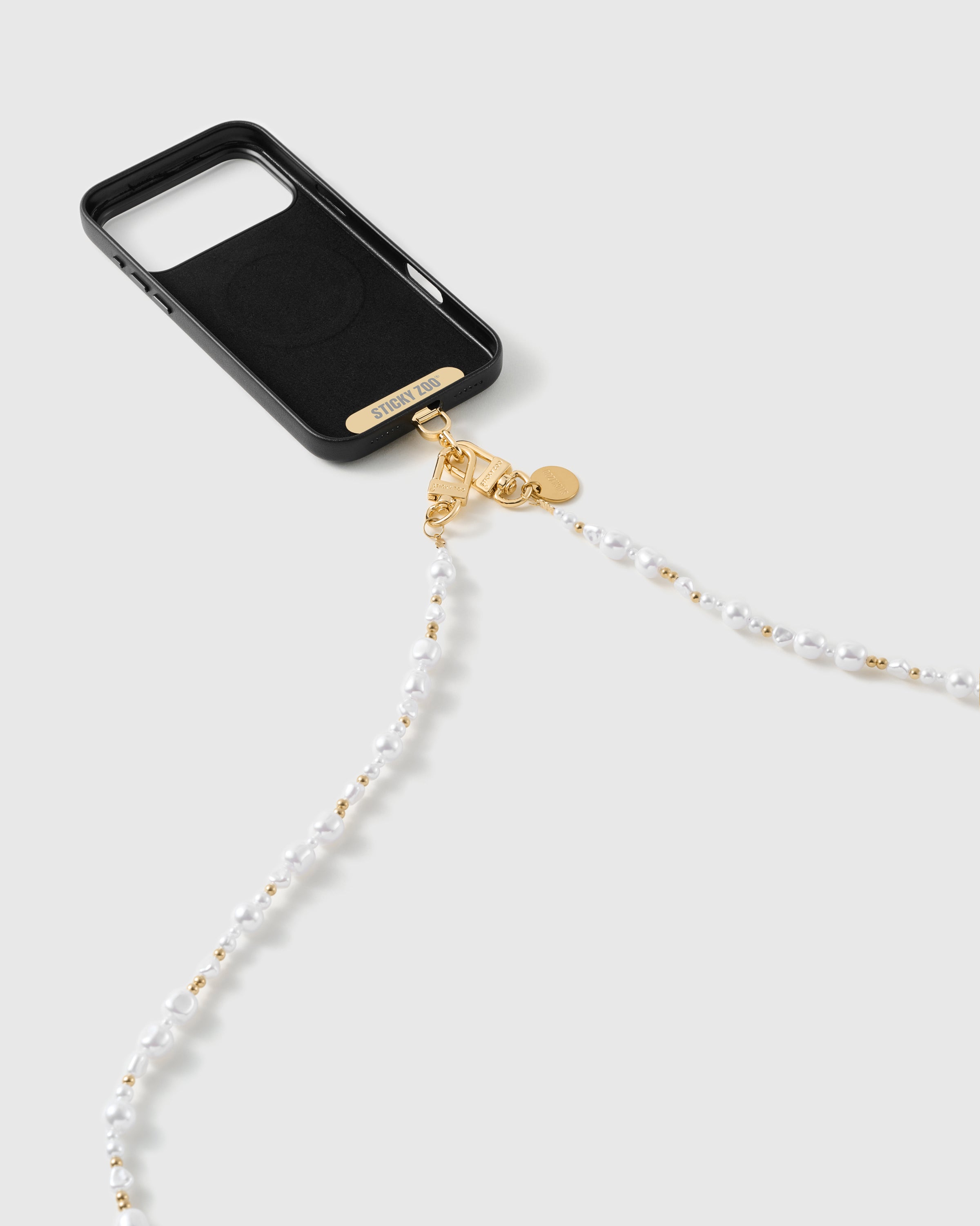 Black phone case with the pearl-and-gold strap draping from its 18K gold-plated dual clasps, gold STICKYZOO phone strap card visible at the case base.