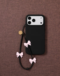 Midnight Bow strap on a black phone case over mauve fabric, pink bows and matte black beads hanging from the cord loop.