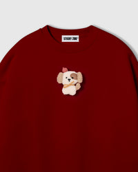 Close-up of hand-stitched plush puppy on red cotton showing cream sherpa body, pink beret, and golden scarf detail