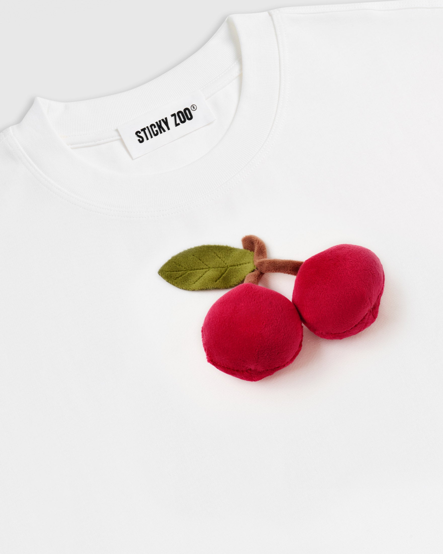 Angled detail of plush cherry pair on STICKYZOO white cotton tee showing dimensional volume and soft texture