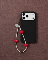 The beaded strap with red hearts and silver seed beads draped beside a black phone case on mauve fabric.