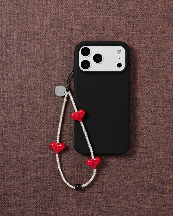 The beaded strap with red hearts and silver seed beads draped beside a black phone case on mauve fabric.