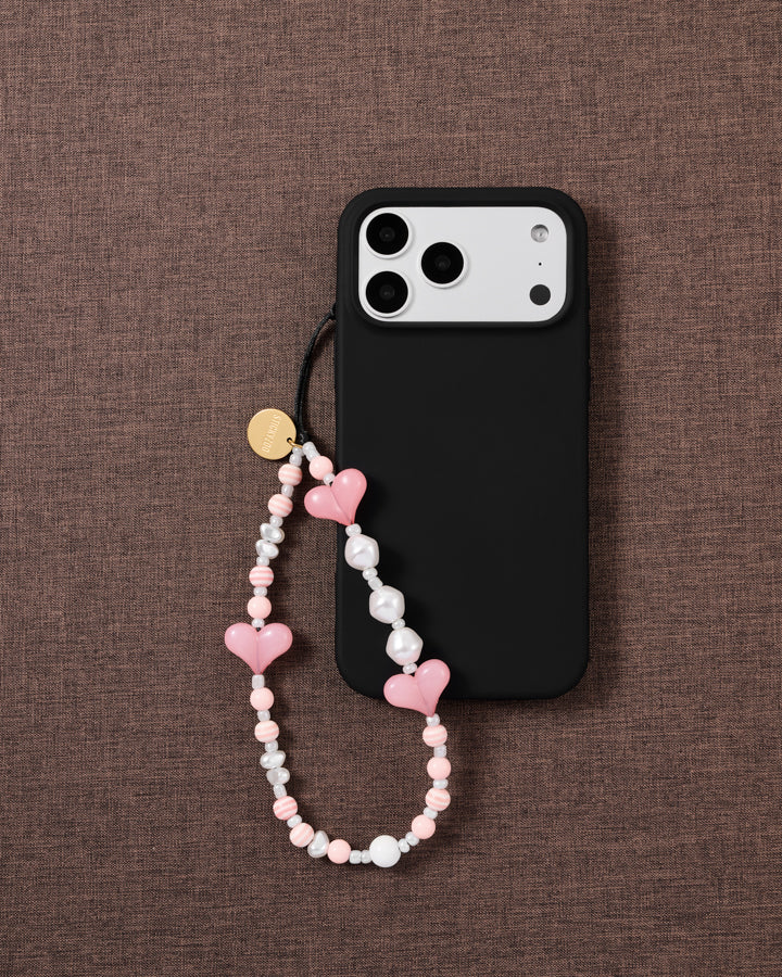 Bubblegum Heart beaded strap looped onto a black phone case on woven brown fabric, candy-pink hearts and pearl-effect beads on show.