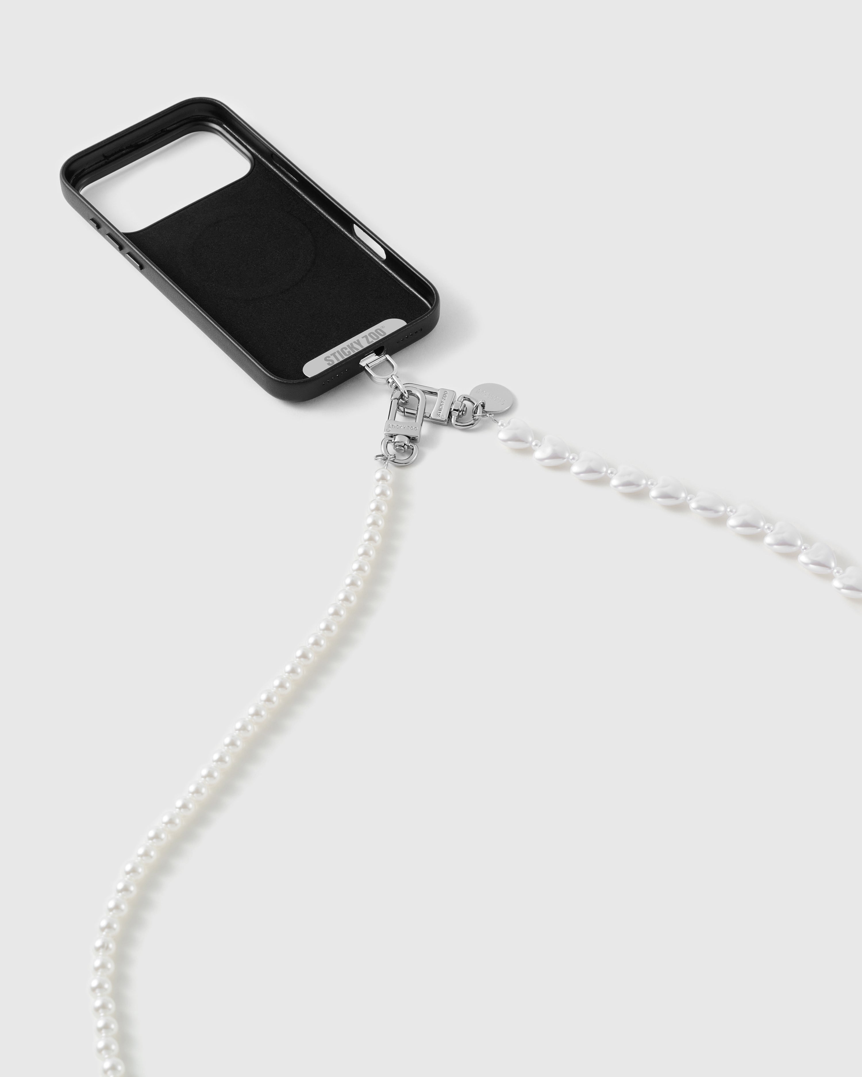 Silver-tone dual clasp linking the faux-pearl and heart strap to a black phone case, pearls trailing across a white studio backdrop.