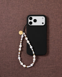 White baroque faux-pearl and gold-bead phone strap looped on a black phone case, styled flat on mauve textured fabric.